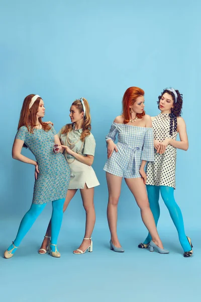 Friends meeting. Full-length portrait of attractive young women posing in stylish clothes against blue studio background. Concept of retro style, fashion, beauty, elegance, 60s, youth. Pin-up style