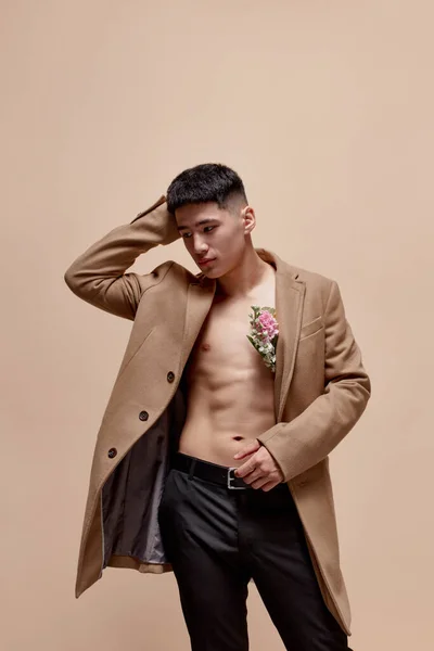 Portrait of handsome young asian guy posing in beige coat on shirtless body with flowers against light brown studio background. Fashion, style, body aesthetics, beauty, mens health, emotions concept