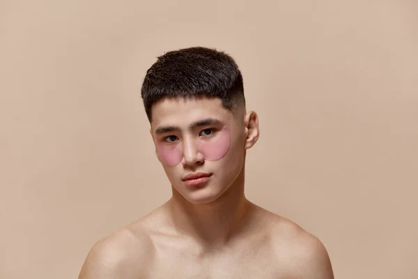 Portrait of handsome young asian guy taking care after skin, applying under eye patches against light brown studio background. Concept of male beauty, skincare, cosmetology, mens health
