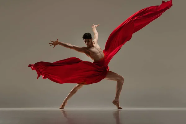 Classic and modern ballet combination. Handsome young male dancer performing with red fabric against grey studio background. Art, classical dance, inspiration, creativity, beauty, choreography concept