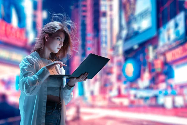 Beautiful young girl standing on lively street and looking on laptop. Working, studying, shopping. Unfocused city background in neon lights. Modern technologies, youth, communication and business