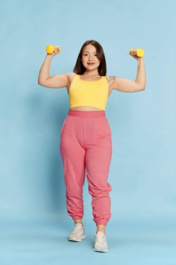 Pembe ve sarı spor kıyafetli Asyalı genç, güzel bir kızın mavi stüdyo arka planında duran dambıllarla tam boy portresi. Duyguların kavramı, yaşam tarzı, gençlik, spor, fitness