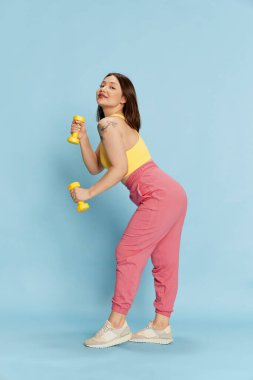 Genç, Asyalı bir kızın rahat spor kıyafetleri içinde uzun bir portresi, spor aletleri ile antrenman, dambıllar mavi stüdyo arka planında. Duyguların kavramı, yaşam tarzı, gençlik, spor, sağlık