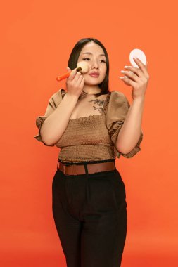 Genç, güzel, şık giyinmiş Asyalı bir kızın portresi, makyaj yapıyor, turuncu stüdyo arka planında yanaklarına allık sürüyor. Duyguların kavramı, yaşam tarzı, gençlik, kozmetoloji, güzellik, moda