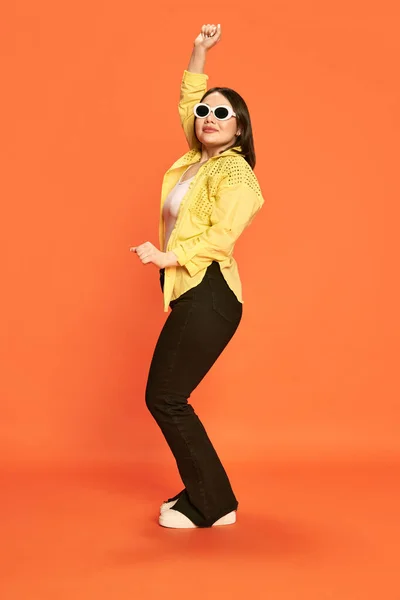 stock image Full-length portrait of beautiful young girl in trendy sunglasses and yellow blouse posing, dancing against orange studio background. Concept of emotions, lifestyle, youth, fashion, fun, celebration