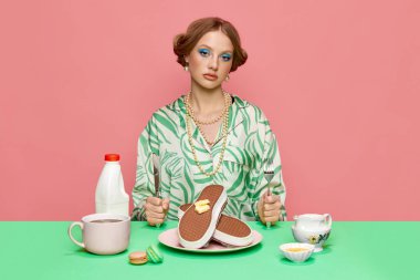 Güzel, şık, genç bir kızın portresi masada oturuyor ve kahvaltıda pembe arka planda tereyağlı waffle yiyor. Yemek ve sanat fotoğrafçılığı. Retro tarzı, yaratıcı görüş konsepti