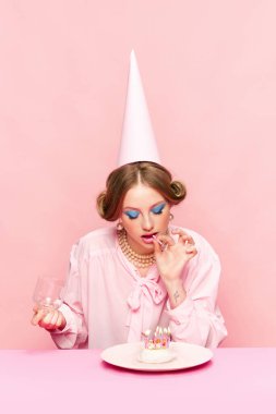 Şık makyajlı bir doğum günü kızı pembe stüdyo geçmişiyle pastaya mum koyuyor. Kutlama. Yemek ve sanat fotoğrafçılığı. Eski tarz, parti, yaratıcı vizyon, hayal gücü, güzellik kavramı