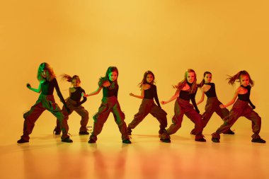 Bir grup yetenekli kız, spor tarzı giyinmiş çocuklar neon ışıkta sarı stüdyo arka planına karşı hip-hop dansı yapıyorlar. Koordineli hareketler. Çocukluk kavramı, hobi, spor yaşam tarzı