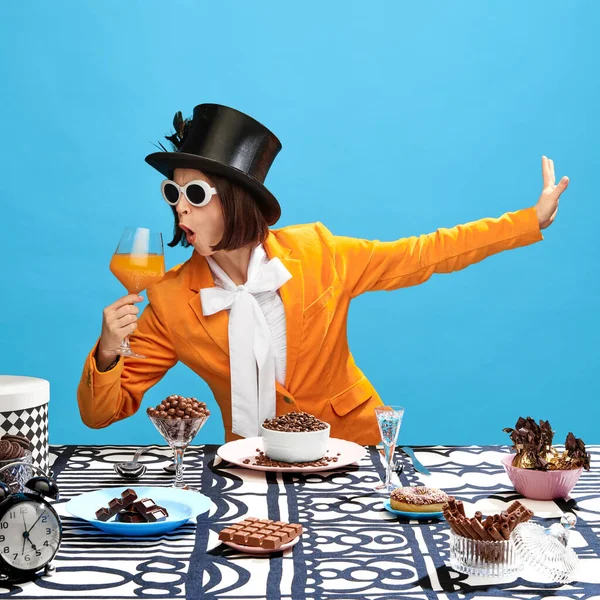 Impressive, eccentric woman in stylish clothes, cylinder and sunglasses, drinking juice over the table full of chocolate. Fairy character. Concept of pop art, creativity, food, inspiration, fashion