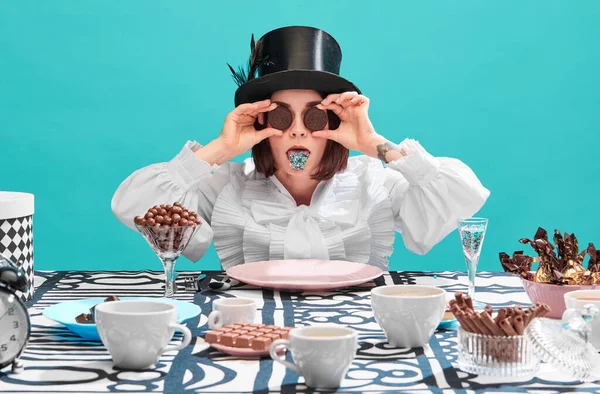 Funny image of woman, pastry chef, sweets lover holding chocolate cookies on eyes, sticking tongue with sparkles over blue background. Concept of pop art, creativity, food, movie character