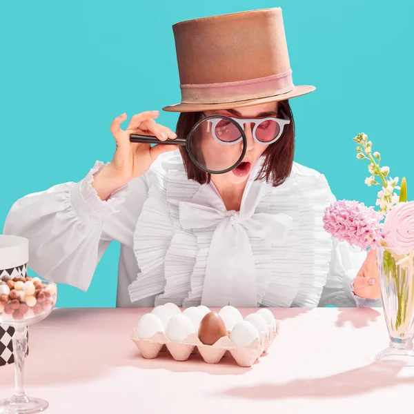 Chocolate egg among ordinary. Woman in cylinder hat looking in magnifying glass with astonished face over blue background. Concept of pop art, creativity, food, inspiration, easter holidays