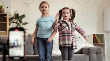 Güzel küçük kızlar, evde dans eden sıradan giysili çocuklar, sosyal medya yayını için cep telefonuyla video kaydı. Kitle iletişim araçları, blog yazma, modern teknolojiler, çocukluk, eğlence kavramı