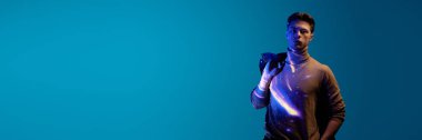 Yakışıklı, ciddi, dijital kosmos 'u olan genç adam neon ışıkta mavi arka planda duran kıyafetlere ve vücuda yansıyor. Modern fotoğrafçılık, sanat, siber punk, tekno, yaratıcılık kavramı. Pankart
