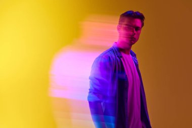 Gündelik giysiler içinde, ciddi bir ifadeyle, karışık neon ışık efektli, değişken sarı arka planda duran genç şık bir adam. Modern fotoğrafçılık, sanat, siber punk, yaratıcılık kavramı