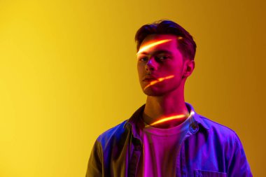 Yüzünde dijital neon filtre yansıması olan günlük tarz kıyafetlerle yakışıklı genç bir adam. Gradyan sarı arka plana karşı duruyor. Modern fotoğrafçılık, sanat, siber punk, tekno, yaratıcılık kavramı