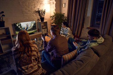 Genç insanlar, oğlanlar ve kızlar, akşamları evdeki kanepede oturan ve konsolda online spor oyunları oynayan arkadaşlar. Neşeli zamanlardı. Arkadaşlık kavramı, eğlence aktivitesi, hobi, eğlence.