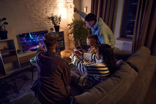 Young people, friends meeting at home in the evening, sitting on couch and playing online sport games, basketball, on console. Concept of friendship, leisure activity, hobby, fun, sport simulator