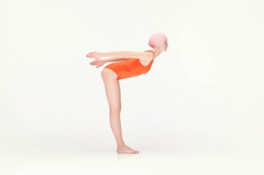 Kırmızı mayo ve pembe yüzme şapkalı genç kız dalış pozisyonu alıyor, beyaz arka planda tek başına eğitim görüyor. Eski tarz, spor, moda, gençlik, klasik. İlan için alanı kopyala