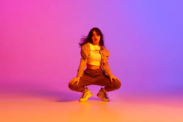 Young expressive girl in sport style clothes dancing modern dance against gradient pink purple background in neon light. Concept of contemporary dance, youth, hobby, action and motion