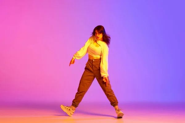 Young female dancer in sport style clothes making performance, training against gradient pink purple background in neon light. Concept of contemporary dance, youth, hobby, action and motion