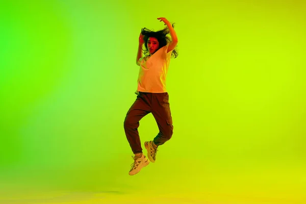 In air. Young girl in sport style clothes dancing, training against gradient green yellow background in neon light. Concept of contemporary dance, youth, hobby, action and motion