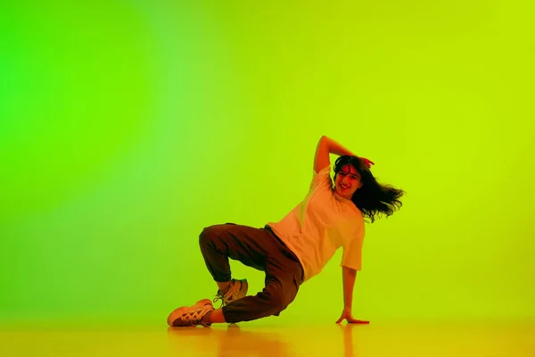 Sliding. Young girl, professional dancer performing hip-hop against gradient green yellow background in neon light. Concept of contemporary dance, youth, hobby, action and motion