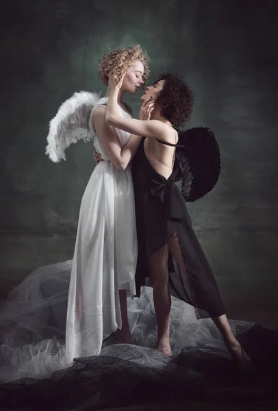 Portrait of two women, angel and demon showing love to each other against dark, green, vintage background. Same-sex marriage. Concept of history, remake, good and bad, creative photography, lgbt