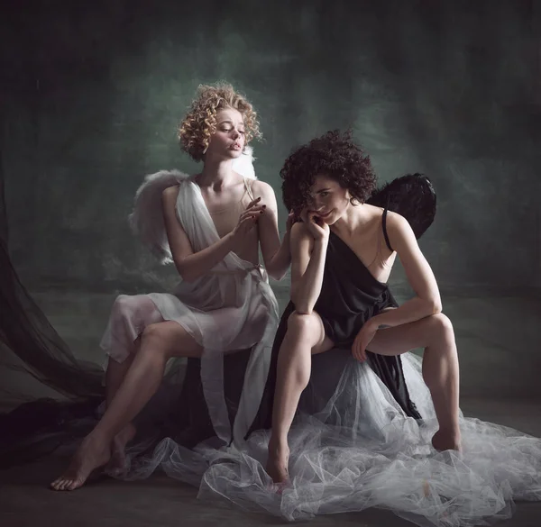 Portrait of two women, queens, angel and demon against dark, green, vintage background. Balance of human being. Concept of history, remake, good and bad, creative photography, fashion