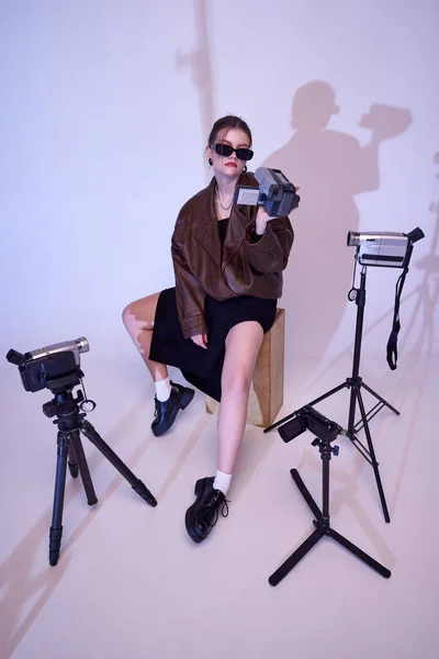 Filming, creating content. Stylish young girl in trendy leather clothes and sunglasses recording videos with retro camera. Concept of fashion, style, retro and vintage, gadgets, beauty, technology