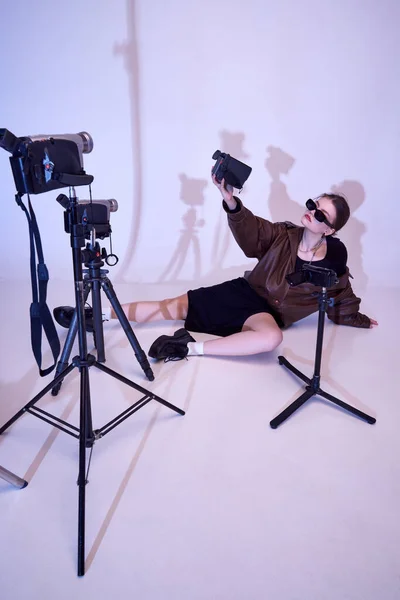 Young girl in stylish leather jacket, dress and trendy sunglasses posing with reto video cameras, filming, making content. Concept of fashion, style, retro and vintage, gadgets, beauty, technology