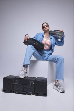 Kot elbiseli genç bir kız. Gri stüdyo arka planında retro müzik çalarıyla oturuyor. Popülerlik. Moda kavramı, 80 'ler, 90' lar tarzı, retro ve vintage, aletler, medya, teknoloji