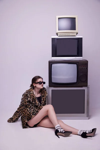 インフルエンサー スタイリッシュな動物のプリントコートとトレトロなテレビセットの近くにポーズをとる流行のサングラスで若い女の子は灰色のスタジオの背景に対して ファッション 80年代 90年代スタイル レトロとヴィンテージ マスメディアの概念 — ストック写真