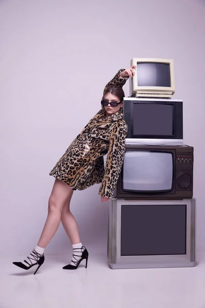 stock image Creative translation. Young girl, influencer in stylish coat and trendy sunglasses posing near retro TV sets against grey background. Concept of fashion, 80s, 90s style, retro and vintage, mass media