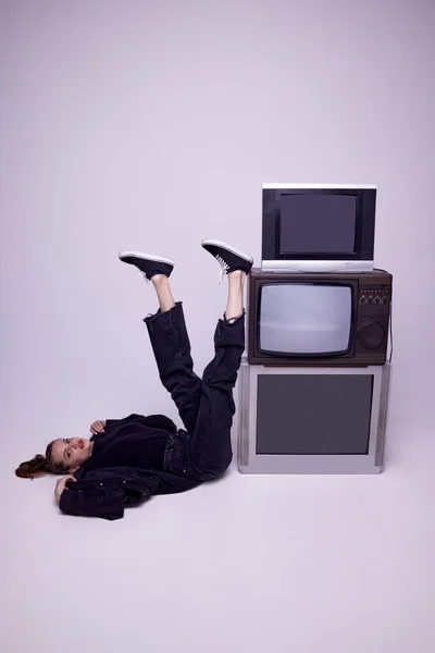 穿着牛仔裤衣服的年轻漂亮的女孩躺在复古电视旁边的地板上 背景是灰色的工作室 电视宣传 时尚概念 80年代 90年代风格 复古风格 大众媒体 — 图库照片