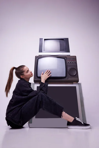 对大众媒体上瘾 穿着黑色衣服的年轻漂亮的女孩坐在复古电视机旁边 背景是灰色的工作室 90年代风格 复古和复古 小玩意 技术概念 — 图库照片
