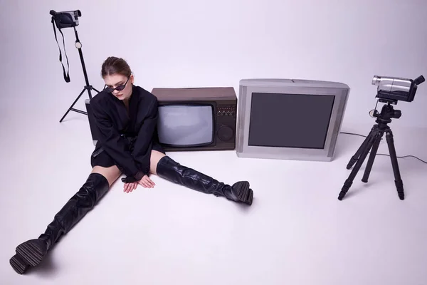 レトロなテレビやカメラの周りの床に座ってスタイリッシュな黒いジャケット 革のブーツやサングラスの若い女の子 ファッション 80年代 90年代スタイル レトロとヴィンテージ ガジェット マスメディアの概念 — ストック写真