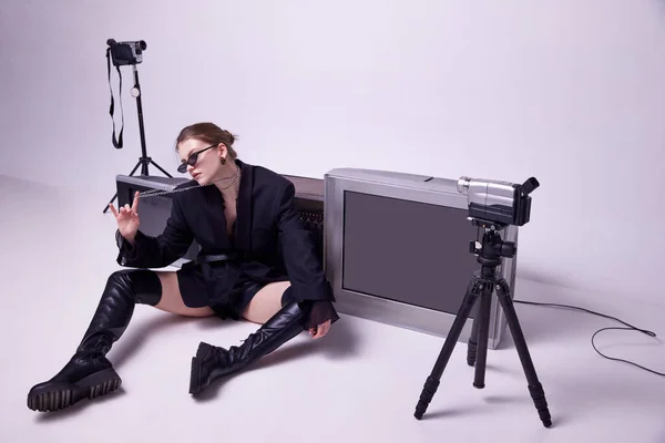 Young girl in stylish black jacket, leather boots and sunglasses sitting on floor around retro TV sets and camera. Concept of fashion, 80s, 90s style, retro and vintage, gadgets, beauty, technology