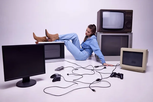 Gamer space. Young girl in jeans clothes sitting on floor around many retro TV sets and playing console. Concept of game, leisure activity, 80s, 90s style, retro and vintage, gadgets