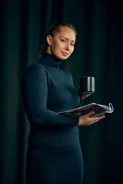 Serious, beautiful, confident woman in elegant clothes standing with tea cup and magazine, looking at camera. Concept of business, fashion, career development, profession, occupation