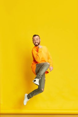 Mutlu, heyecanlı, sıradan giysiler içinde, parlak sarı arka planda zıplayan sakallı bir adamın tam boy stüdyo portresi. İnsan duyguları kavramı, yaşam tarzı, yüz ifadesi, başarı, kutlama. Ad