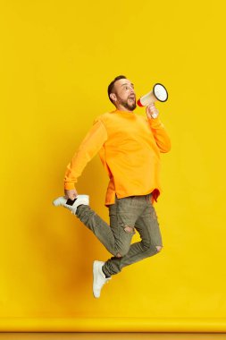 Sakallı bir adamın tam boy portresi, etkili, atlayan ve bağıran megafonla canlı sarı stüdyo arkaplanı. İnsan duyguları, yaşam tarzı, yüz ifadesi kavramı. Ad