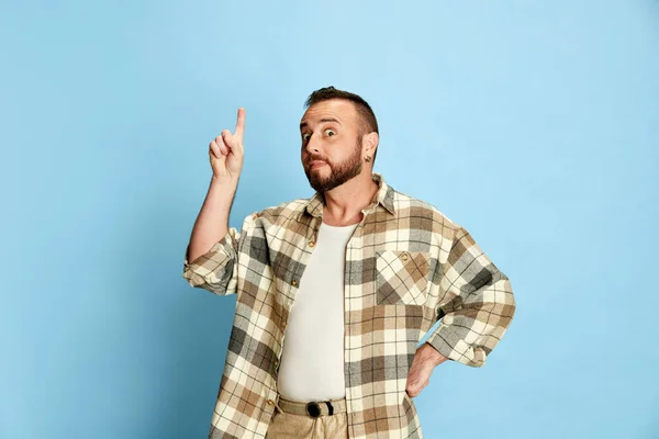 カジュアルな服を着た髭の男の肖像画 チェックしたシャツが指を上げる 青いスタジオの背景に対するアイデアを表現しています 人間の感情 ライフスタイル 表情の概念 — ストック写真
