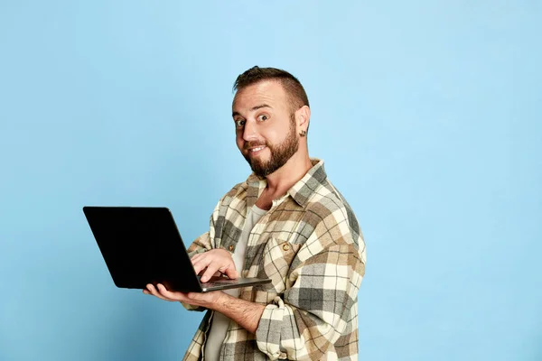 Portrait of smiling, positive, bearded man in checkered shirt holding tablet and working against blue studio background. Concept of human emotions, lifestyle, facial expression, freelance job. Ad