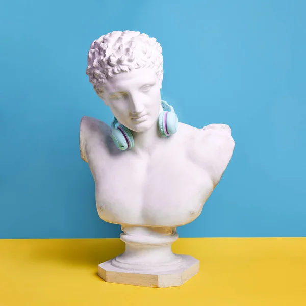 Antique statue bust wearing blue headphones against blue background. Music lifestyle, technologies, youth culture. Concept of creativity, modernity and vintage, antique art. Inspiration, imagination