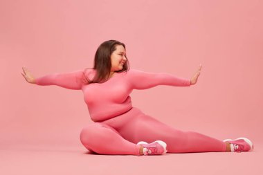 İyi olmak ve kendini sevmek. Pembe spor giyim üzerine çalışan şişman bir kadın. Pembe stüdyo geçmişine karşı esneme hareketleri yapıyor. Spor anlayışı, vücut pozitifliği, kilo kaybı, vücut ve sağlık hizmetleri