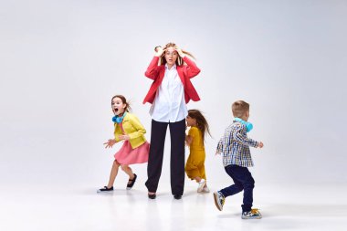 Yorgun genç bir kadının portresi, annesinin başını tutması küçük gürültücü çocukların gri stüdyo arka planında oynaması. Aile kavramı, annelik, çocukluk, moda, yaşam tarzı