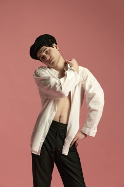 Pembe stüdyo arka planında poz veren şık beyaz bluzlu, pantolonlu ve siyah bereli genç yakışıklı bir adam portresi. Paris tarzı, klasik görünüş. Erkek modası, stil, sanat, güzellik kavramı