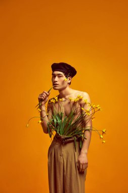Rahatlık ve tarz. Siyah bereli, klasik bej pantolonlu, sarı stüdyo arka planında çiçeklerle üstsüz poz veren yakışıklı bir adamın portresi. Erkek modası, sanat, güzellik, trend kavramı
