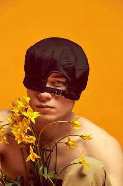 Dört gözle bekliyorum. Sarı arka planda siyah bereli üstsüz poz veren genç adam. Çiçeklerle poz ver. Erkekler moda, tarz, sanat, güzellik, trendler, ilham