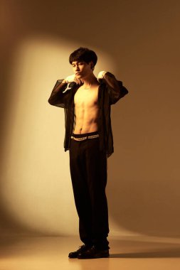 Stüdyonun arka planında çıplak vücut üzerinde şeffaf gömlekle poz veren yakışıklı bir gencin tam boy portresi. Erkek modası, tarzı, sanatı, güzelliği, eğilimleri
